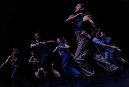 Sur cette image, il y a 7 danseuses et danseurs en train d'effectuer tous ensemble le même mouvement de danse, jambes gauche croisée devant la jambe droite, avec les mains en contact au niveau de leur poitrine.
