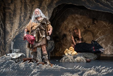© Théâtre de Carouge / Cédric Vincensini Un homme préhistorique à l'air hargneux, un gros morceau de viande saignante à la main, regarde devant lui, pendant qu'un autre homme préhistorique, derrière, lui dépèce un bœuf mort.