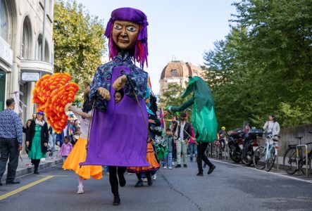 Grande marionnette du personnage zuza dans la rue avec des acteurs et des actrices qui la suivent et des personnes du public autour.