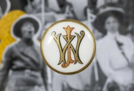 Un bouton en céramique au fond blanc avec des lettres dorés et à la bordure dorée est disposé sur une image en noir et blanc et légèrement floue représentant des femmes.