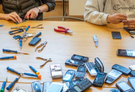 Assis à une table, des personnes dont on ne voit pas la tête sont en train de démonter des téléphones. Sur la table s'étalent des téléphones de toutes sortes, des tournevis et des pinces