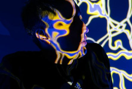 ©Lisa Frisco L'image montre des fils lumineux jaunes et violets sur un fond bleu nuit. Ces fils lumineux sont projetés sur un jeune homme que l'on devine plus que l'on voit. L'image a un effet un peu mystérieux et magnétique. Ces fils sont en réalité des projections de VJing.