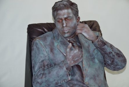 Homme habillé en costume avec son corps et vêtements peints en gris argenté. Il fait face à la caméra et est assis sur une chaise en cuir devant un mur gris clair. Son regard est au sol à droite alors que sa main gauche tient le col du costume prêt de son cou et sa main droite tient la partie proche de sa poitrine.