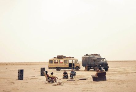 Un desert, deux voitures et plusieurs personnes assises sur des chaises et un canapé devant les voitures. Il y a également deux grosses enceintes pour du son.