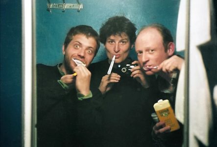 © Riverboom Trois personnes se prennent en photo dans un miroir tout en se brossant les dents, souriantes et décontractées, dans un espace exigu à l’éclairage tamisé.