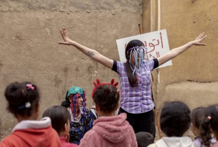 © Les Filles du Nil Performance de rue animée par deux femmes costumées devant un groupe de jeunes filles, dans une ruelle. L’une lève les bras, l’autre est assise. Une pancarte en arabe est visible en arrière-plan. Ambiance ludique et engagée.
