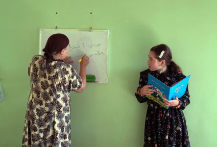 une adulte et une jeune fille, se tiennent devant un tableau blanc fixé à un mur vert. L’adulte écrit en arabe pendant que la jeune fille lit un livre illustré