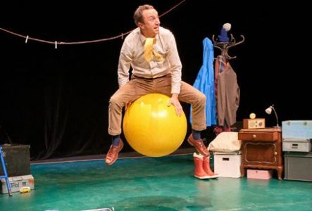 Une scène de théâtre est aménagée avec de nombreux objets du quotidien. Au centre, une personne est assise à califourchon sur un grand ballon jaune de gymnastique. La personne porte une chemise claire, un pantalon beige et des chaussures marron.
À gauche de la scène se trouve une échelle bleue. Un fil à linge est tendu en hauteur d’un côté à l’autre de l’espace, avec plusieurs pinces à linge accrochées. Derrière la personne, un porte-manteau supporte plusieurs vêtements colorés.
À droite, un…