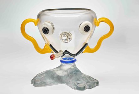 Œuvre de Buddy Di Rosa. Il s'agit d'une coupe en verre transparent sur lequel un visage a été créé en volume, avec une cigarette à la bouche.