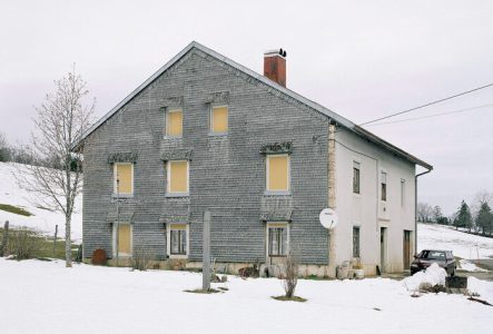Photographie d’une maison jurassienne grise au volets jaunes fermées, dans la neige.