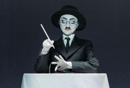 Une personne habillée en mime est assis à une table avec une nappe blanche sur fond noir. Elle porte un costume noir, des gants blancs et une cravate noire.