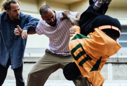 Photographie prise dans un espace extérieur ou semi-ouvert aux couleurs claires. Trois personnes occupent le centre de l’image et semblent participer à une performance de danse ou de mouvement.
À gauche, une personne porte une longue chemise bleu foncé et un pantalon sombre. Les bras sont pliés et légèrement écartés du corps dans une posture dynamique.
Au centre, une personne vêtue d’une chemise claire à fines rayures verticales et d’un pantalon beige est penchée vers l’avant. Un bras est posé…