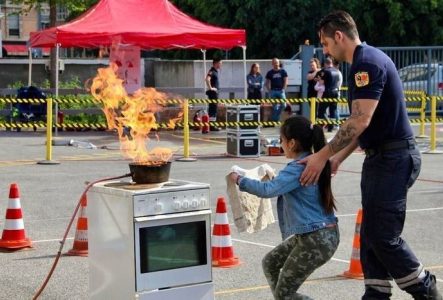 Lors d'une démonstration de sécurité sur un parking, une enfant, guidée par un sapeur-pompier, s'approche d'une casserole en flammes sur une cuisinière, un linge à la main pour étouffer le feu.