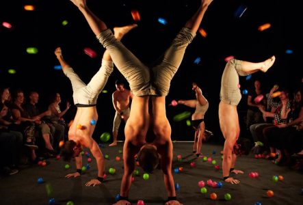 Des acrobates sont en équilibre sur les mains pendant que le public leur jette des balles de couleur dessus