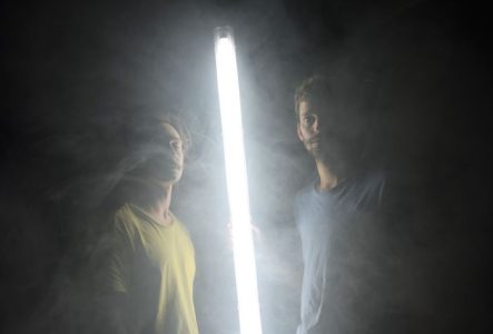 Deux comédiens sont dans le noir, éclairés uniquement par un tube lumineux placé entre eux.