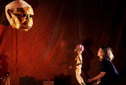 Une comédienne et une marionnette de table font face a une grosse tête d'un personnage préhistorique en marionnette à fils.