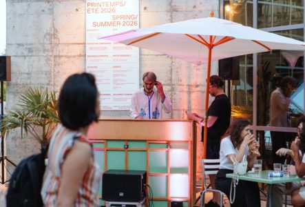 DJ derrière une console lors d’un événement en terrasse, sous un grand parasol blanc, tandis que des visiteurs discutent et prennent un verre autour de tables dans un espace contemporain éclairé par une lumière chaude.