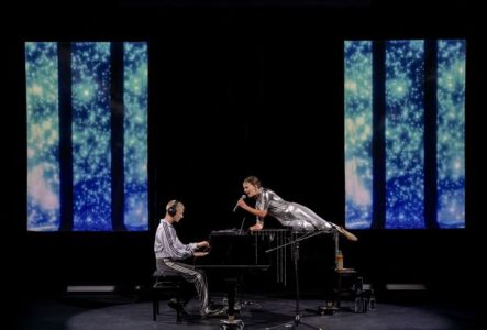 Un homme joue du piano, un casque sur les oreilles. Devant lui, une femme est allongée, un micro à la main. Tous deux portent des vêtements argentés. Deux panneaux lumineux sont placés de chaque côté de la scène