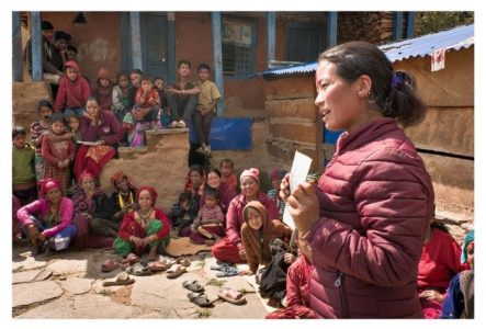 Une séance de sensibilisation de l’organisation partenaire Sahakarmi au Népal en 2025. On voit, de côté, une femme népalaise qui tient une fiche dans ses mains. Elle est en train de parler à une assemblée communautaire dont on voit quelques membres, femmes, hommes et enfants, assis dans un cercle autour d'elle au sol et sur des escaliers d'un bâtiment au fond de l'image.