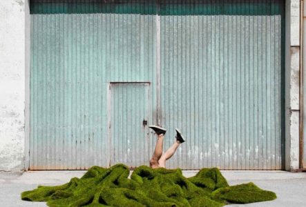couverture verte de laquelle ressortent deux jambes de deux comédiens différents au devant d'une entrée de garage