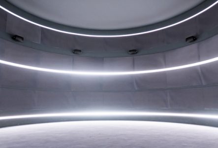 Espace dans l'exposition Afrosonica. Vision panoramique d'un espace circulaire sombre. Trois lignes circulaires de néon, du sol au plafond structure le tout. L'image est dans les ton sombres et gris et les néons ressortent très lumineux.