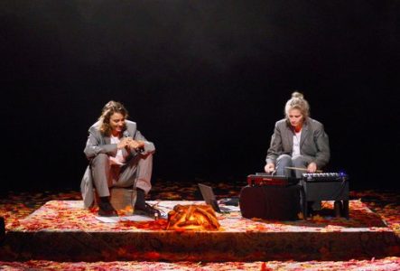 Deux comédiennes sont assises sur scène. L'une avec un micro dans les mains, l'autre bat le rythme avec des baguettes. Le sol est recouvert de feuilles mortes rouges et oranges.