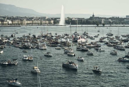 Une foule impressionnante de bateaux est rassemblée sur le lac Léman lors de l'événement On The Water Geneva Festival. En arrière-plan, on observe le Jet d'eau de Genève, avec une vue sur la ville, la cathédrale et un aperçu du Salève