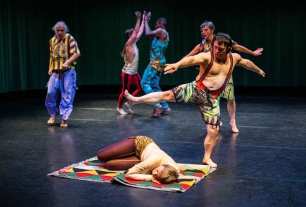 Sur scène, une danseuse est allongée au sol, sur un tapis coloré. Derrière elle, cinq autres interprètes dansent debout.
