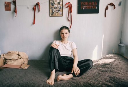 Une jeune fille est assise sur son lit, appuyée contre le mur décoré de sa chambre. Un cadre, rempli de photographies personnelles avec la mention 2025, un panneau où y est écrit en lettres rouges Yobana Pusnya (en ukrainien) et des rubans rouges sont suspendus au mur.